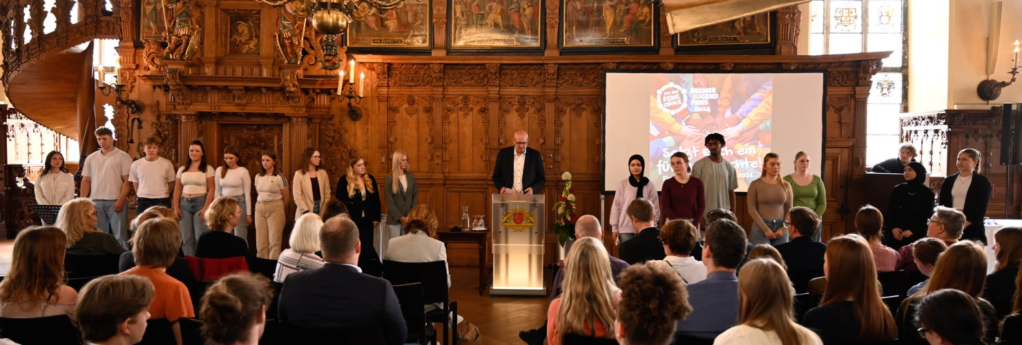 Auf dem Bild befindet sich eine formelle Veranstaltung in einem Raum mit historischer Holzarbeit und einem prachtvollen Kronleuchter. Eine Gruppe von jungen Erwachsenen steht auf einer Holzbühne vorne, wobei ein Mann am Podium spricht und auf eine digitale Projektion hinter ihm hinweist. Das Publikum, bestehend aus Erwachsenen verschiedener Altersgruppen, schaut aufmerksam zu.