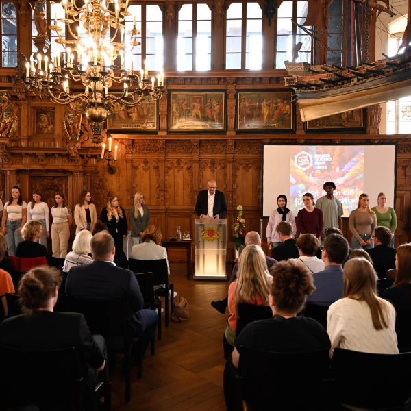 Auf dem Bild befindet sich eine formelle Veranstaltung in einem Raum mit historischer Holzarbeit und einem prachtvollen Kronleuchter. Eine Gruppe von jungen Erwachsenen steht auf einer Holzbühne vorne, wobei ein Mann am Podium spricht und auf eine digitale Projektion hinter ihm hinweist. Das Publikum, bestehend aus Erwachsenen verschiedener Altersgruppen, schaut aufmerksam zu.  
