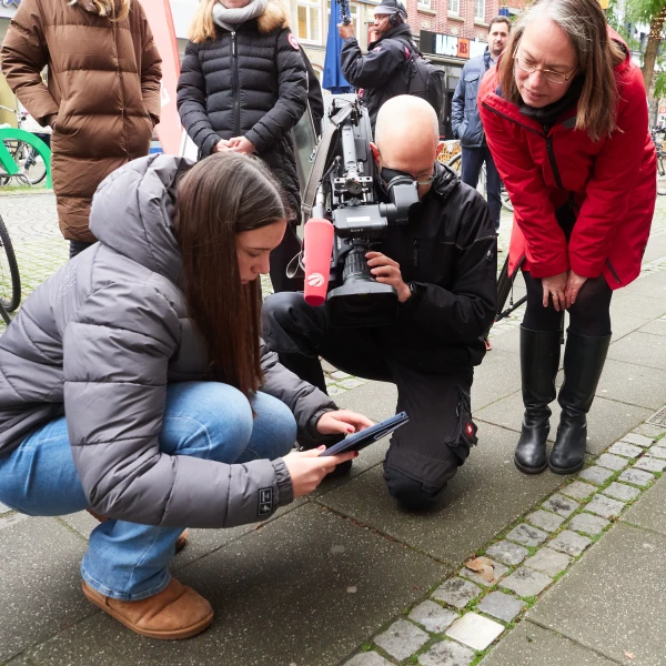 Auf dem Bild ist eine Gruppe von Menschen zu sehen, die sich auf einem gepflasterten Gehweg befindet. Eine Frau in einem braunen Stiefel und einer grauen Jacke ist gehockt und bedient ein Tablet, während ein Mann mit einer Kamera, die an einem Gestell befestigt ist, neben ihr kniet. Eine andere Frau in einer roten Jacke beobachtet interessiert das Geschehen, umgeben von weiteren Personen, die im Hintergrund stehen.  