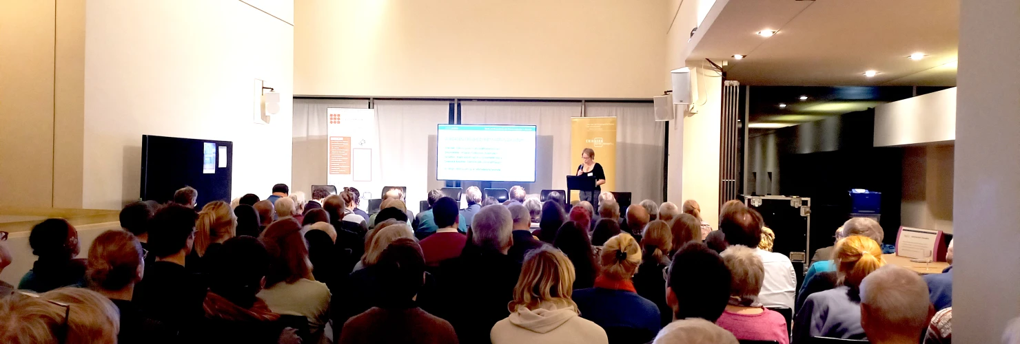 Ein voll besetzter Vortragssaal: Eine Person steht vorne am Rednerpult und präsentiert eine Folie auf einer Leinwand, während das Publikum aufmerksam zuhört.  