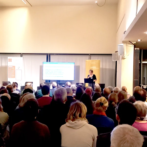 Ein voll besetzter Vortragssaal: Eine Person steht vorne am Rednerpult und präsentiert eine Folie auf einer Leinwand, während das Publikum aufmerksam zuhört.  