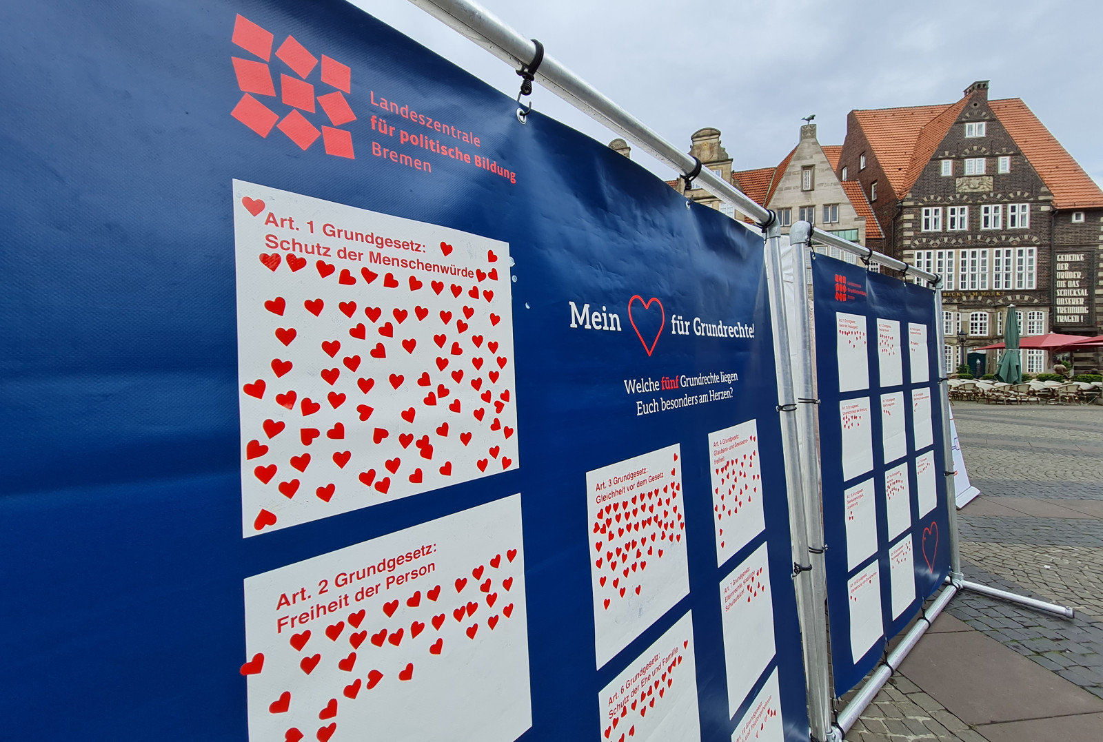 Das Bild zeigt eine Ausstellung im Freien mit zwei blauen Informationswänden, die eng zusammenstehen und grafisch gestaltete Plakate über Grundrechte tragen. Auf einem der sichtbaren Plakate steht "Art. 1 Grundgesetz: Schutz der Menschenwürde" umgeben von vielen roten Herzen, und ein weiteres erwähnt "Art. 2 Grundgesetz: Freiheit der Person". Im Hintergrund ist der Marktplatz von Bremen zu erkennen.  