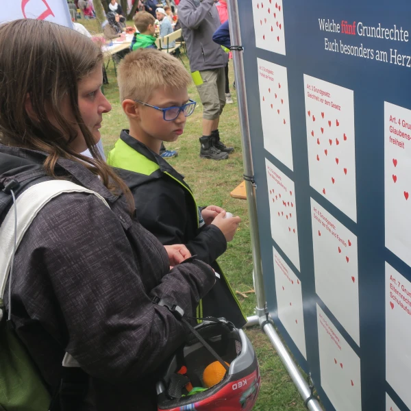 Auf diesem Bild sind eine junge Frau und ein Junge, die vor einem großen Informationsstand stehen, auf welchem die Grundrechte des Grundgesetzes aufgedruckt sind. Der Stand ist draußen auf einer Veranstaltung aufgestellt, wobei etliche rote Herz-Aufkleber auf der blauen Fläche angebracht sind, die zeigen, welche Grundrechte den Menschen wichtig sind. Die Frau trägt einen Rucksack und hat langes Haar, während der Junge eine Brille trägt.  