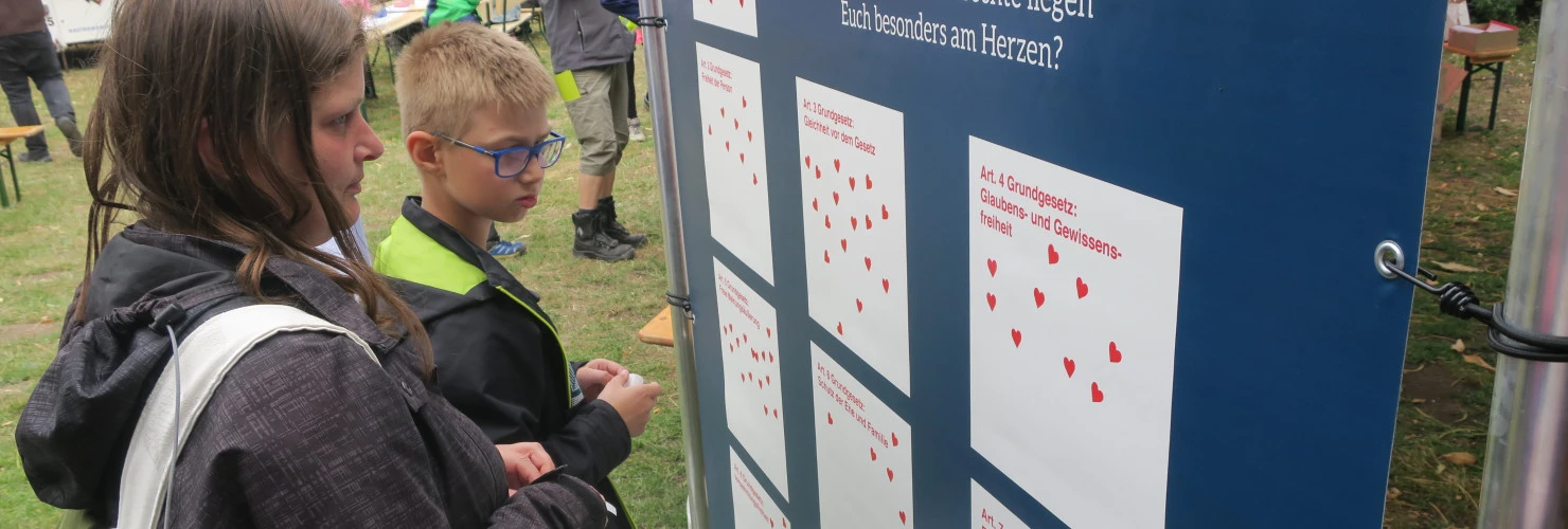 Auf diesem Bild sind eine junge Frau und ein Junge, die vor einem großen Informationsstand stehen, auf welchem die Grundrechte des Grundgesetzes aufgedruckt sind. Der Stand ist draußen auf einer Veranstaltung aufgestellt, wobei etliche rote Herz-Aufkleber auf der blauen Fläche angebracht sind, die zeigen, welche Grundrechte den Menschen wichtig sind. Die Frau trägt einen Rucksack und hat langes Haar, während der Junge eine Brille trägt.  