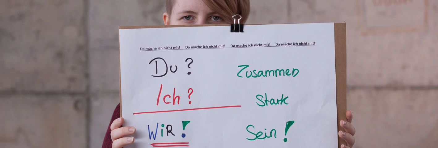 Auf dem Bild ist eine junge Frau mit kurzen rotbraunen Haaren, die ein großes Plakat vor ihren Körper und ihren Mund hält. Auf dem weißen Plakat stehen in bunten Farben die Worte „Du? Ich? Wir.“ gefolgt von „Zusammen Stark Sein!“, wobei jedes Wort in einer anderen Farbe (rot, grün, blau und nochmals grün) geschrieben ist. Der Hintergrund hinter der Frau ist eine graue Betonwand.  