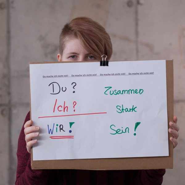 Auf dem Bild ist eine junge Frau mit kurzen rotbraunen Haaren, die ein großes Plakat vor ihren Körper und ihren Mund hält. Auf dem weißen Plakat stehen in bunten Farben die Worte „Du? Ich? Wir.“ gefolgt von „Zusammen Stark Sein!“, wobei jedes Wort in einer anderen Farbe (rot, grün, blau und nochmals grün) geschrieben ist. Der Hintergrund hinter der Frau ist eine graue Betonwand.  