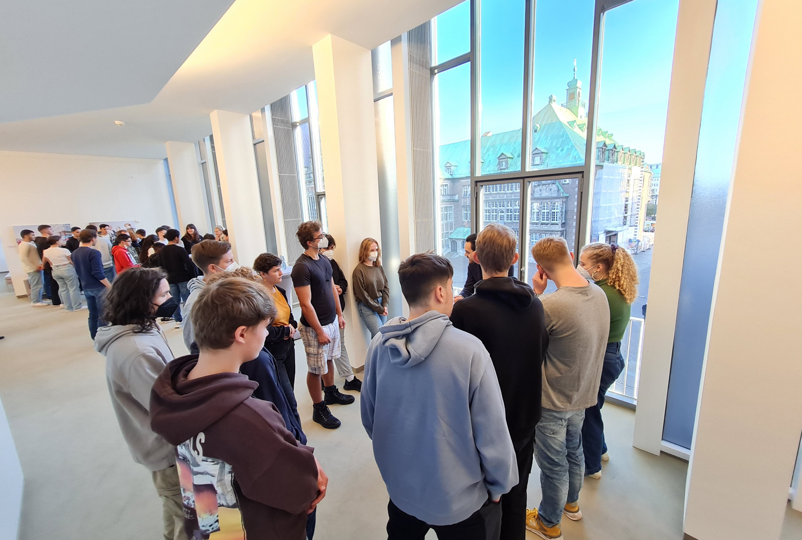 Das Bild zeigt eine Gruppe von jungen Erwachsenen, die im Inneren der Bremischen Bürgerschaft stehen und durch bodentiefe Fenster nach draußen schauen. Sie blicken auf das Bremer Rathaus. Die Atmosphäre wirkt ruhig und gesellig, während die Personen in ungezwungener Kleidung wie Jeans und Hoodies miteinander kommunizieren.  