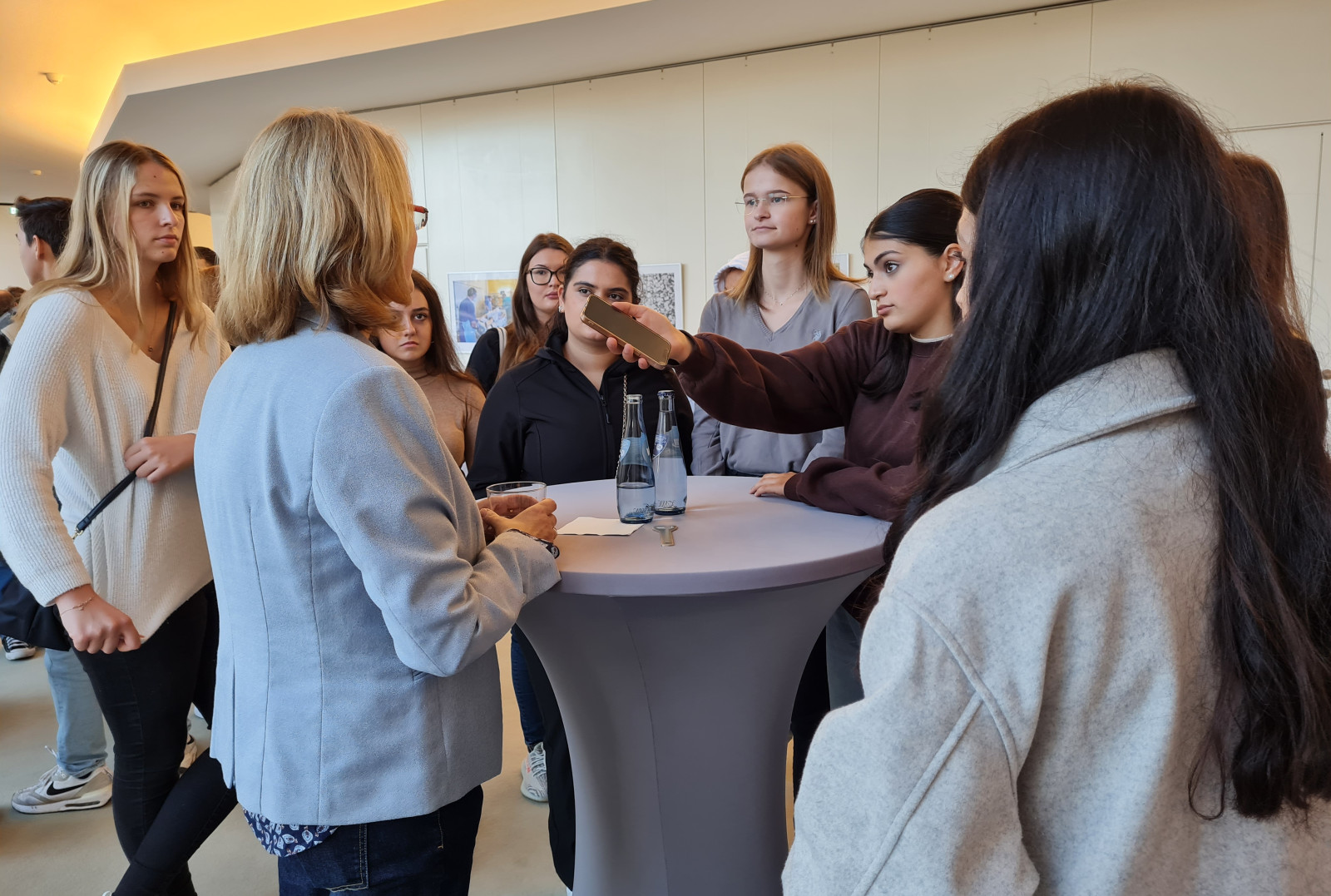 In einem Innenraum der Bremischen Bürgerschaft stehen mehrere Personen, hauptsächlich junge Frauen, um einen Stehtisch versammelt. Sie unterhalten sich engagiert mit einer Abgeordneten. Eine Schülerin hält ein Diktiergerät, während andere aufmerksam zuhören.   