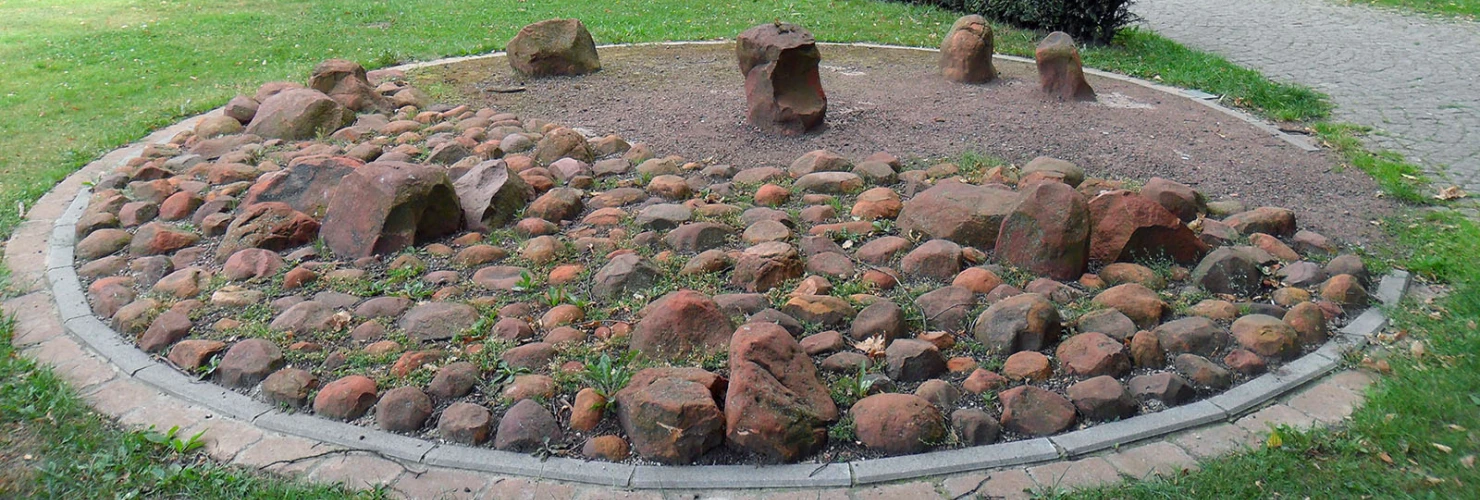 Das Bild zeigt einen Park mit einem großen Elefanten-Steindenkmal in einer Naturumgebung. Im Vordergrund befindet sich ein kreisförmiges Beet, welches mit vielen kleinen und großen Steinen gefüllt ist. Der Hintergrund wird von grünen Bäumen und einem gepflasterten Weg, der durch den Park führt, geprägt.