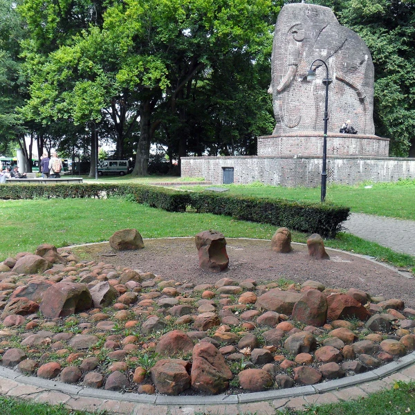 Das Bild zeigt einen Park mit einem großen Elefanten-Steindenkmal in einer Naturumgebung. Im Vordergrund befindet sich ein kreisförmiges Beet, welches mit vielen kleinen und großen Steinen gefüllt ist. Der Hintergrund wird von grünen Bäumen und einem gepflasterten Weg, der durch den Park führt, geprägt.  