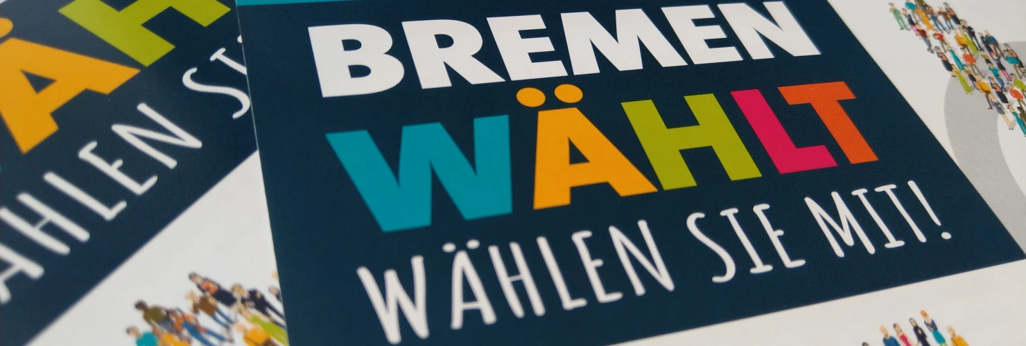 Auf dem Bild sind mehrere bunte Flyer zu sehen, die für die Wahl in Bremen am 26. September 2021 werben. Die Überschrift auf den Flyern lautet "BREMEN WÄHLT" in großen, fetten Buchstaben. Unterhalb der Überschrift findet sich der Zusatz "Wählen Sie mit!", begleitet von Illustrationen zahlreicher kleiner Menschenfiguren, die Vielfalt und Teilnahme symbolisieren.  