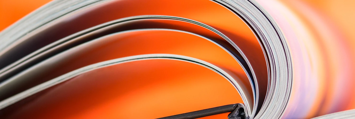 Das Bild zeigt eine Nahaufnahme von den Seiten eines aufgeschlagenen Buches. Die Seiten sind geschwungen und formen sanfte Wellen. Im Hintergrund ist eine unscharfe orange Fläche zu erkennen, die einen kontrastreichen, farbigen Hintergrund bietet.  