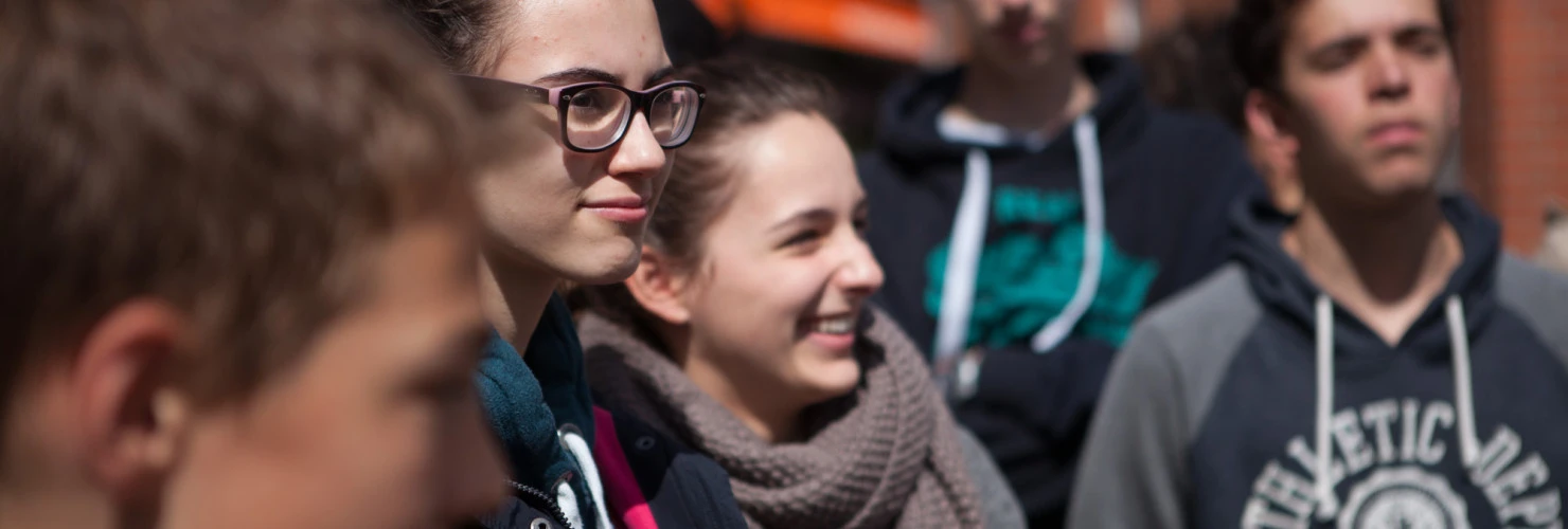 Eine Gruppe junger Menschen steht im Freien und schaut in verschiedenen Richtungen, wobei der Fokus auf drei Mädchen in der Bildmitte liegt, die lächeln und klare Brillen tragen. Im Vordergrund ist das Gesicht eines Jungen unscharf, und im Hintergrund steht ein weiterer Junge mit einem Hoodie. Die Jugendlichen scheinen sich in einer lebhaften Unterhaltung oder einer Aktivität zu befinden.  