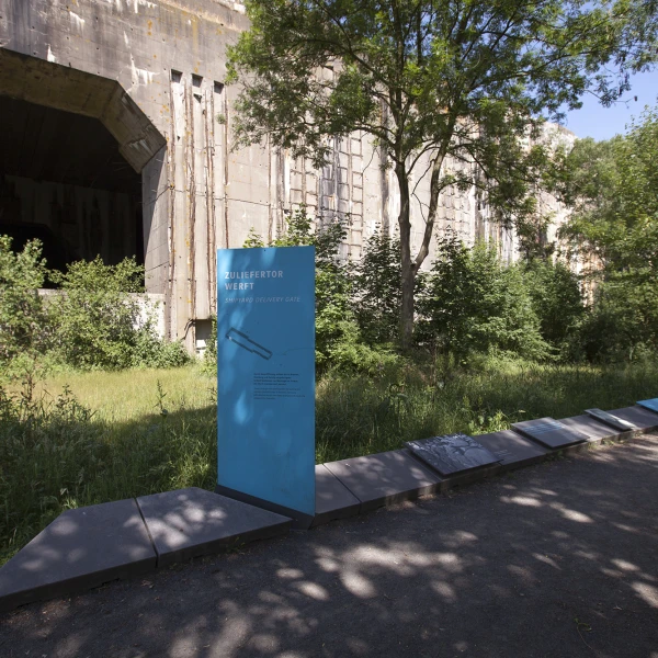 Eine blaue Informationstafel am Rundweg des Denkort Bunker Valentin. Im Hintergrund ist der Bunker selbst. Das Bild zeigt das alte Betongebäude mit einem offenen, dunklen Eingang, umgeben von üppigem Grün.   