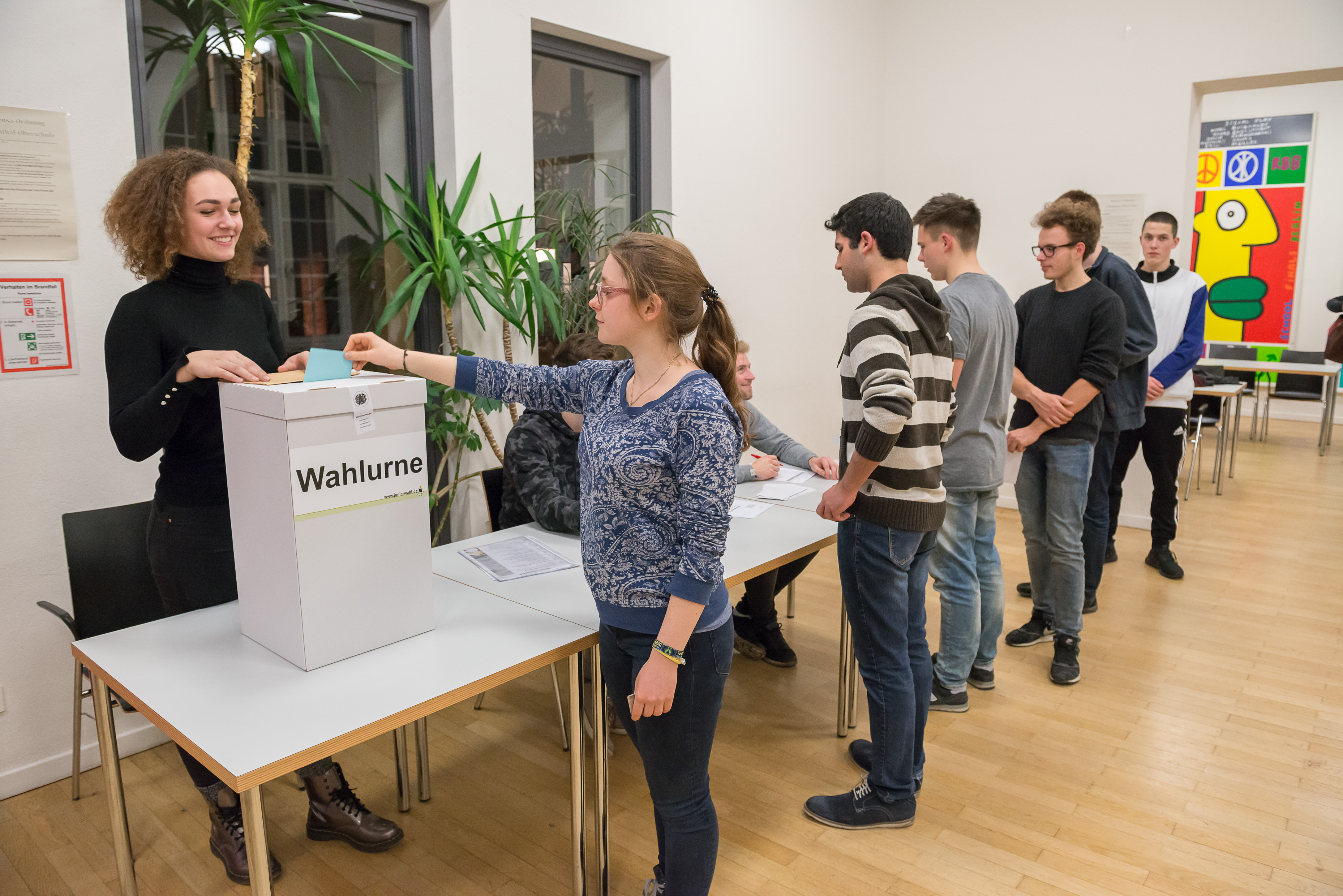 Eine Gruppe junger Menschen stellt sich an der Wahlurne an. Im Vordergrund wirft eine junge Frau ihren Wahlschein in diese Wahlurne ein.   