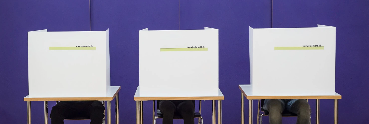 Drei Wähler:innen sitzen in einem Wahllokal hinter jeweils einem Sichtschutz.  
