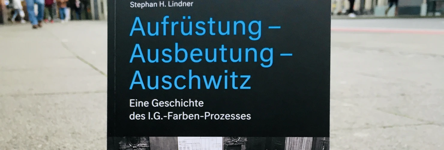 Ein Bild des Buches: "Aufrüstung-Ausbeutung-Auschwitz. Eine Geschichte des I.G. -Farben-Prozesses" von Stephan H. Lindner mit dem Hintergrund einer städtischen Straße.  