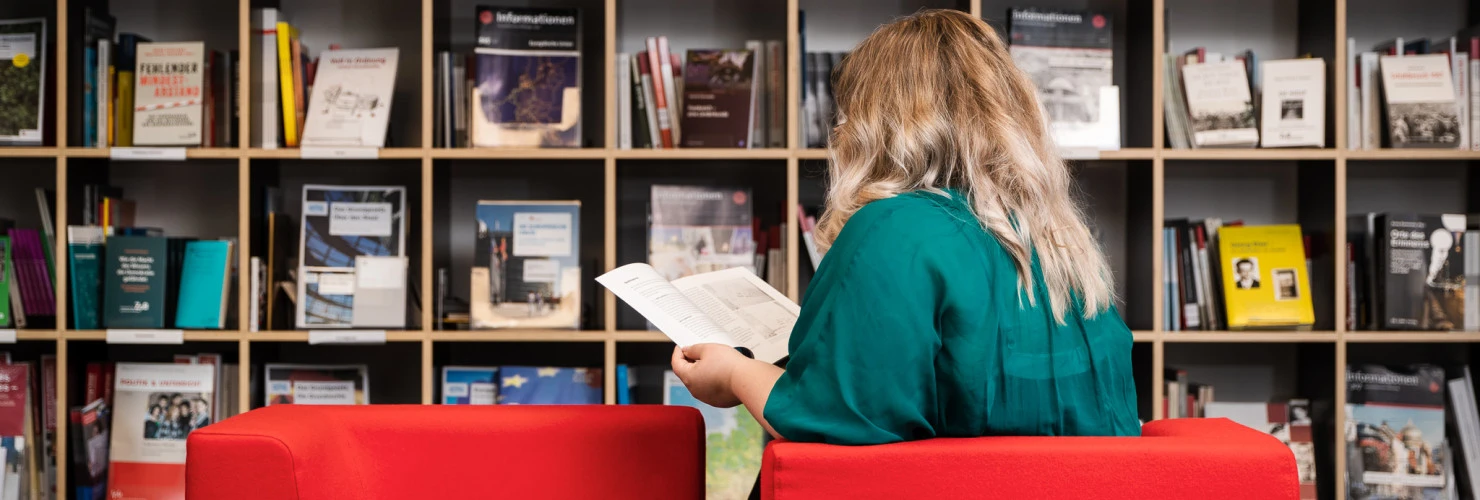 Eine Frau sitzt mit dem Rücken zur Kamera vor einem Bücherschrank und ließt ein Buch.