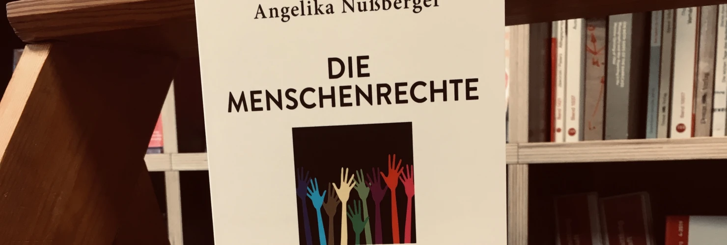 Ein Bild des Buches "Die Menschenrechte. Geschichte, Philosophie, Konflikte" von Angelika Nußberger mit dem Hintergrund aus Bücherregalen.    