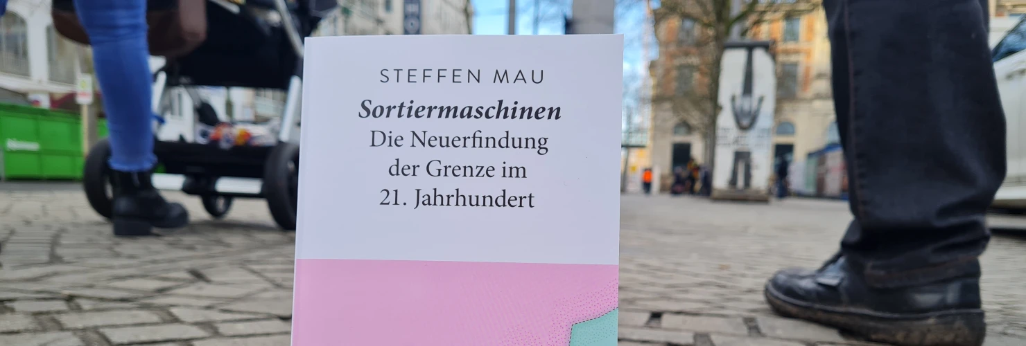 Ein Bild des Buches "Sortiermaschinen-Die Neuerfindung der Grenze im 21. Jahrhundert" von Steffen Mau mit einem Hintergrund einer städtischen Straße.  