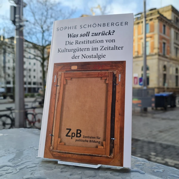 Ein BIld des Buches: " Was soll zurück? Die Restitution von Kulturgütern im Zeitalter der Nostalgie" von Sophie Schönberger mit dem Hintergrund einer städtischen Straße.  