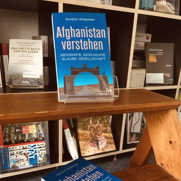 Das Cover des Buches "Afghanistan verstehen. Geographie, Geschichte, Glaube, Gesellschaft" mit einem Foto vom Bogen von Qala-e-Bost. Im Hintergrund sind Regale mit verschiedenen Büchern zu sehen.  