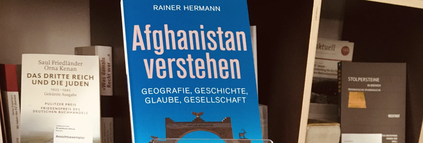 Das Cover des Buches "Afghanistan verstehen. Geographie, Geschichte, Glaube, Gesellschaft" mit einem Foto vom Bogen von Qala-e-Bost. Im Hintergrund sind Regale mit verschiedenen Büchern zu sehen.  