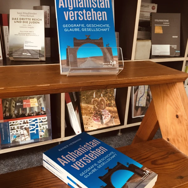 Das Cover des Buches "Afghanistan verstehen. Geographie, Geschichte, Glaube, Gesellschaft" mit einem Foto vom Bogen von Qala-e-Bost. Im Hintergrund sind Regale mit verschiedenen Büchern zu sehen.  
