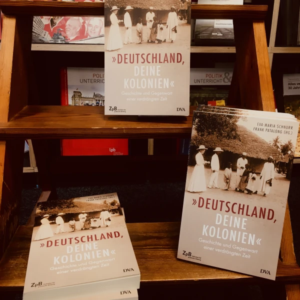 Ein Buchcover " Deutschland, deine Kolonien - Geschichte und Gegenwart einer verdrängten Zeit" mit der Abbildung eines Schwarzweißfotos von Menschen, die vor traditionellen afrikanischen Hütten stehen und weiß gekleidet sind. Zu sehen sind auch drei Kinde  