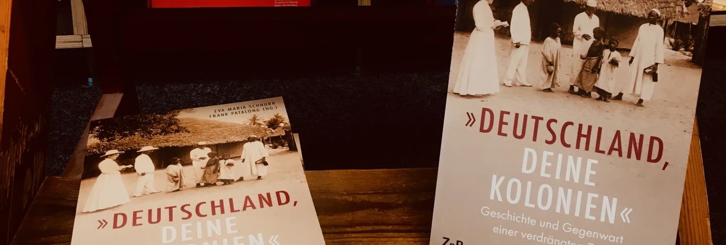 Ein Buchcover " Deutschland, deine Kolonien - Geschichte und Gegenwart einer verdrängten Zeit" mit der Abbildung eines Schwarzweißfotos von Menschen, die vor traditionellen afrikanischen Hütten stehen und weiß gekleidet sind. Zu sehen sind auch drei Kinde