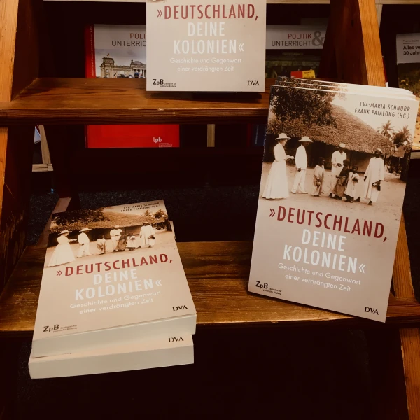 Ein Buchcover " Deutschland, deine Kolonien - Geschichte und Gegenwart einer verdrängten Zeit" mit der Abbildung eines Schwarzweißfotos von Menschen, die vor traditionellen afrikanischen Hütten stehen und weiß gekleidet sind. Zu sehen sind auch drei Kinde  