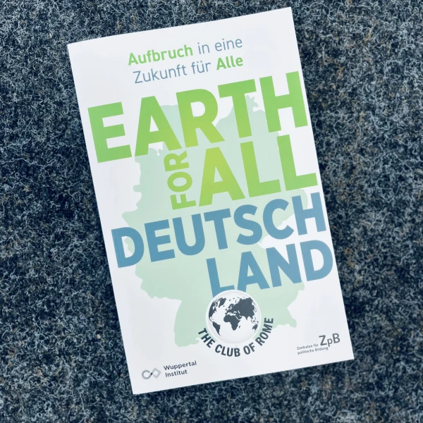 Das Bild zeigt ein Buch mit dem Titel "EARTH FOR ALL DEUTSCHLAND" auf einem grauen Hintergrund. Der obere Teil des Buchumschlags ist in Weiß gehalten, darauf steht "Aufbruch in eine Zukunft für Alle". Im unteren Bereich des Umschlags ist eine einfache, grüne Illustration Deutschlands zu erkennen, darüber prangt in großen, grünen Buchstaben der Haupttitel, sowie der Untertitel "Deutschland" in blauer Schrift.  