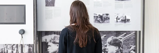 Eine Person steht mit dem Rücken zur Kamera und betrachtet eine Ausstellungswand, die mit historischen Fotografien und Textpanelen bestückt ist. Die Person ist in einem dunklen, legeren Outfit gekleidet, bestehend aus einer Bluse und weiten Hosen, und trägt auffällige, rot-grün gestreifte Schuhe. Der Ausstellungsraum hat helle Wände und einen grauen Boden, was für einen neutralen Hintergrund sorgt.  