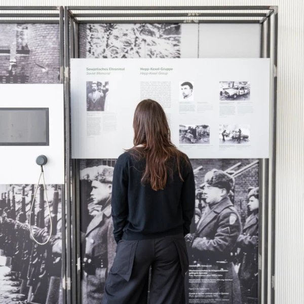 Eine Person steht mit dem Rücken zur Kamera und betrachtet eine Ausstellungswand, die mit historischen Fotografien und Textpanelen bestückt ist. Die Person ist in einem dunklen, legeren Outfit gekleidet, bestehend aus einer Bluse und weiten Hosen, und trägt auffällige, rot-grün gestreifte Schuhe. Der Ausstellungsraum hat helle Wände und einen grauen Boden, was für einen neutralen Hintergrund sorgt.  