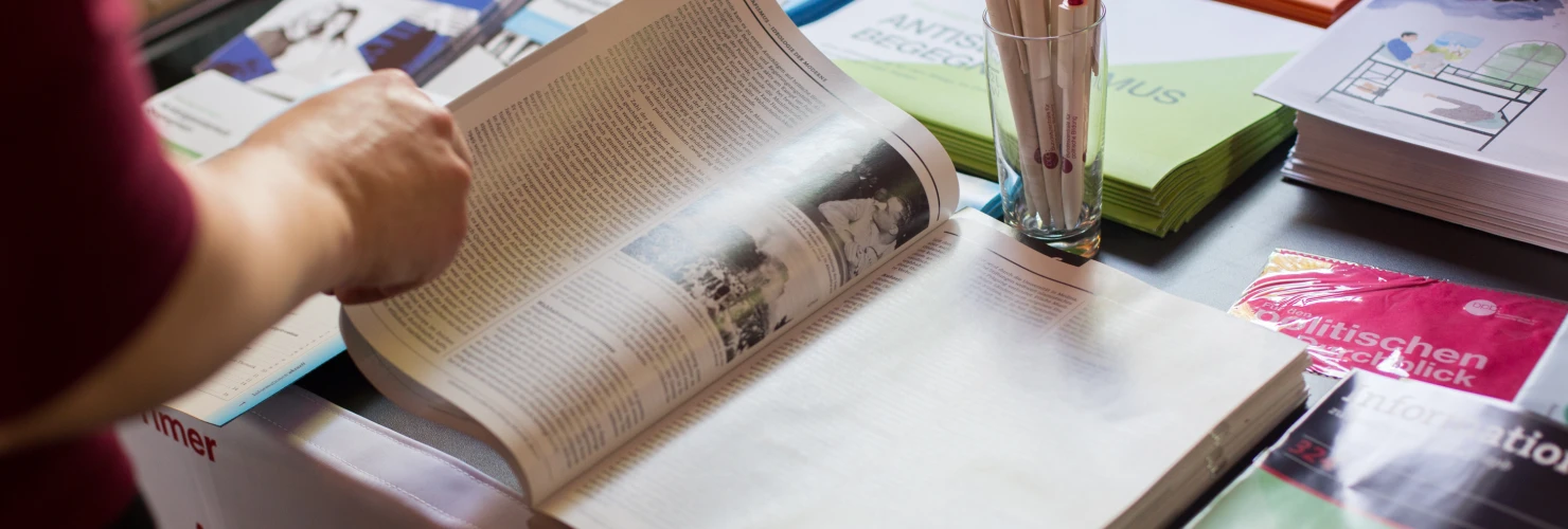 Auf dem Bild ist eine Person, die an einem Informationsstand mit verschiedenen Büchern und Broschüren steht und gerade eine aufgeschlagene Zeitschrift durchblättert. Die Person ist nicht vollständig abgebildet, sondern nur der rechte Arm. Der Stand trägt ein Schild mit der Aufschrift "Websites und Zeitschriften". Im Vordergrund auf dem Tisch steht ein Glas  gefüllt mit mehreren Kugelschreibern.  