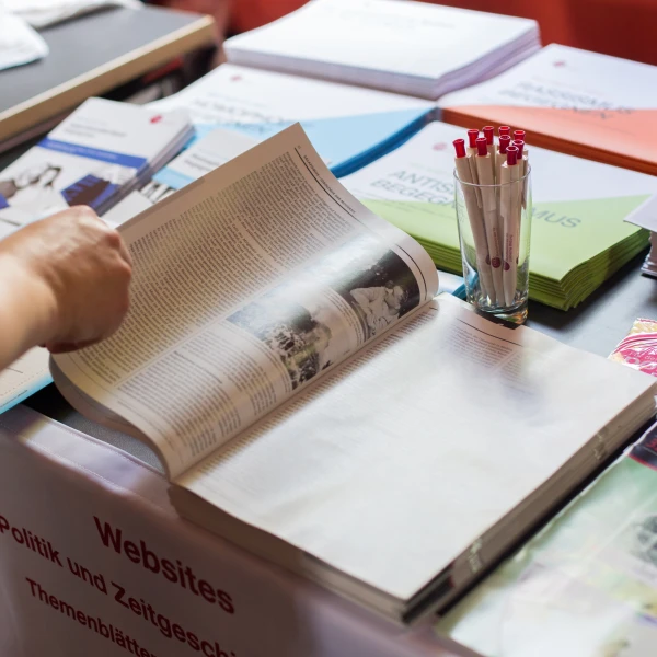 Auf dem Bild ist eine Person, die an einem Informationsstand mit verschiedenen Büchern und Broschüren steht und gerade eine aufgeschlagene Zeitschrift durchblättert. Die Person ist nicht vollständig abgebildet, sondern nur der rechte Arm. Der Stand trägt ein Schild mit der Aufschrift "Websites und Zeitschriften". Im Vordergrund auf dem Tisch steht ein Glas  gefüllt mit mehreren Kugelschreibern.  