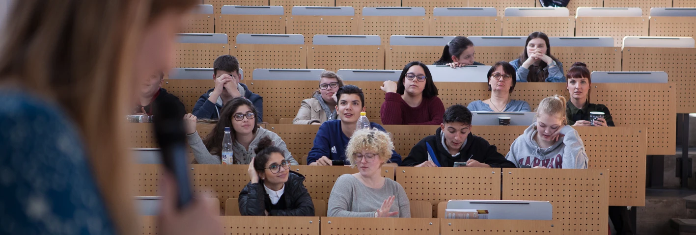 Es ist ein Hörsaal mit mehreren Reihen von Holzsitzen, auf denen junge Menschen verschiedenen Alters und Geschlechts sitzen und nach vorne blicken. Manche Reihen sein frei. Eine junge Frau mit langen Haaren steht im Vordergrund an einem Pult und spricht, während sie auf ein Mikrofon nahe an ihrem Mund hält. Sie ist auf dem Bild verschwommen. Die Menschen in den Stuhlreihen wirken ruhig und hören aufmerksam zu.  