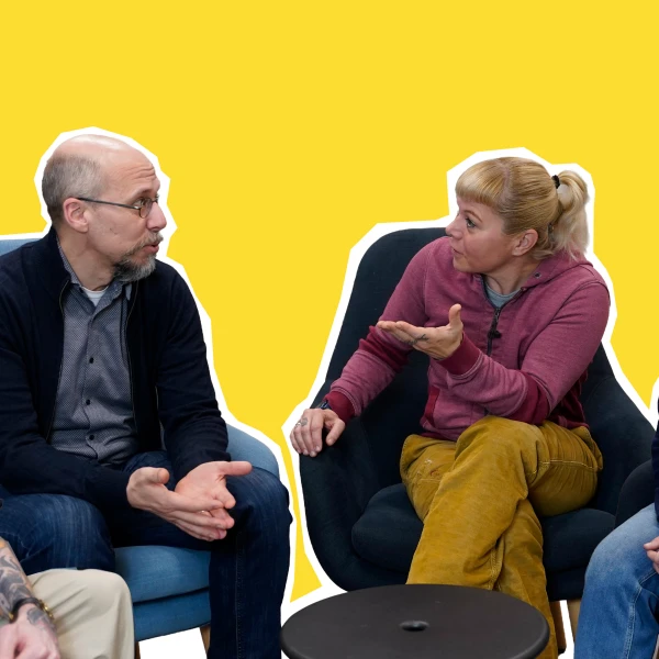 Auf dem Bild ist eine Gruppe von fünf Erwachsenen in einer entspannten Diskussionssituation in einem Innenraum zu sehen. Zwei Personen in der Mitte, eine Frau und ein Mann, sind in eine intensivere Unterhaltung vertieft und scheinen etwas zu diskutieren, wobei die Frau gestikulierend spricht und der Mann aufmerksam zuhört.   