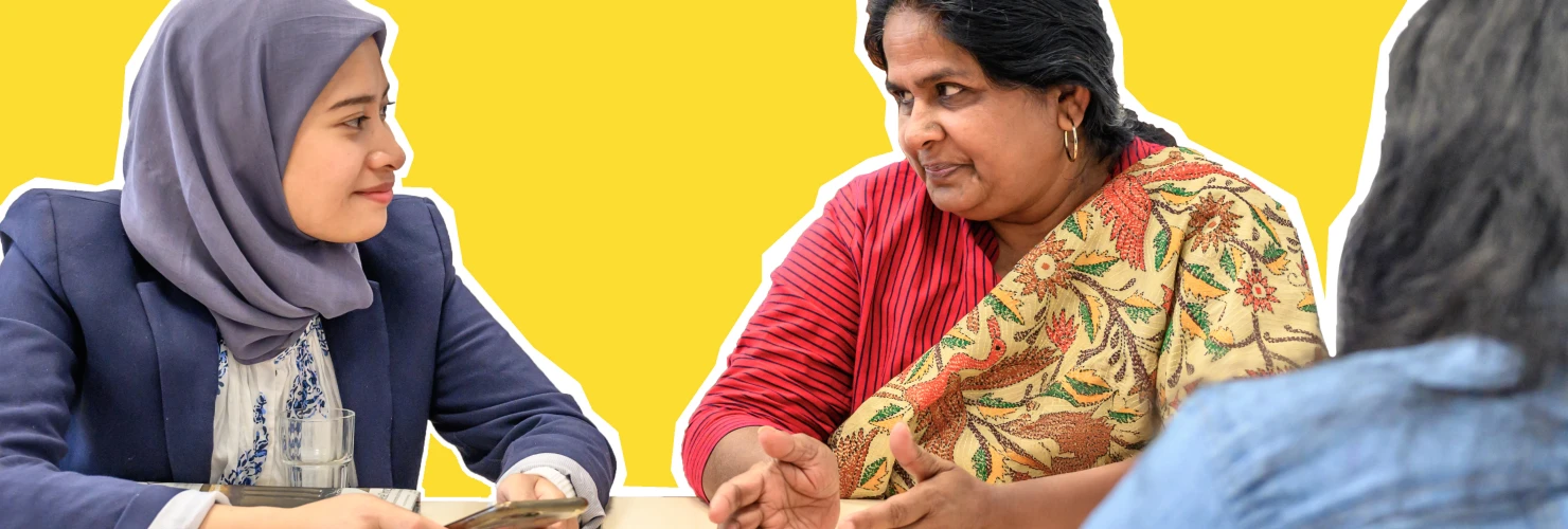 Auf dem Bild sitzen drei Frauen um einen weißen Tisch, vor einem gelben Hintergrund. Eine junge Frau mit einem blauen Hijab und einem hellblauen Jackett hält ein Handy und unterhält sich mit einer älteren Frau in einem roten Oberteil und einer gemusterten Jacke, die ihr aufmerksam zuhört. Eine dritte Frau, deren Rücken zur Kamera zeigt, trägt ein dunkelblaues Top.  