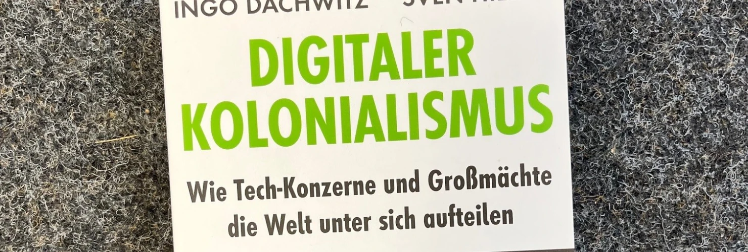 Auf dem Bild ist das Cover eines Buches zu sehen, das auf einem grauen Teppichboden liegt. Der Titel des Buches lautet "Digitaler Kolonialismus" und stammt von Ingo Dachwitz und Sven Hilbig. Das Cover zeigt eine Weltkarte in verschiedenen Grüntönen mit verschwommenen Konturen, die von digitalen Netzwerklinien und Punkten überzogen ist.  