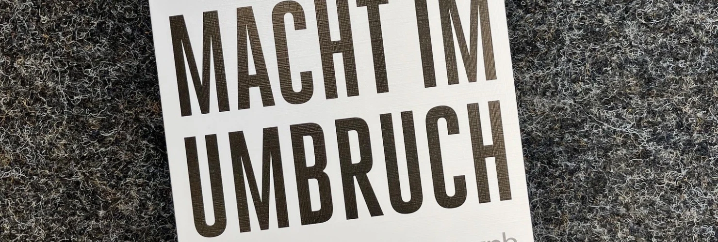Auf dem Bild ist ein Buch mit dem Titel "Macht im Umbruch" zu sehen, das auf einer grauen Oberfläche liegt. Der Autor des Buches, Herfried Münkler, wird auf dem Cover oben in kleineren schwarzen Buchstaben erwähnt. Das Cover zeigt große, weiße und schwarze Buchstaben und enthält den Untertitel „Deutschlands Rolle in Europa und die Herausforderungen des 21. Jahrhunderts“ in kleinerer schwarzer Schrift.  