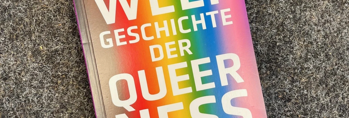 Auf dem Bild liegt ein Buch mit dem Titel "Weltgeschichte der Queerness" auf einem grauen Untergrund. Das Buchcover zeigt einen farbenfrohen Regenbogeneffekt, der von links unten nach rechts oben verläuft. Der Name des Autors "Dirk Wicke" und der Name des Verlags "zpb" sind ebenfalls auf dem Cover zu sehen.  
