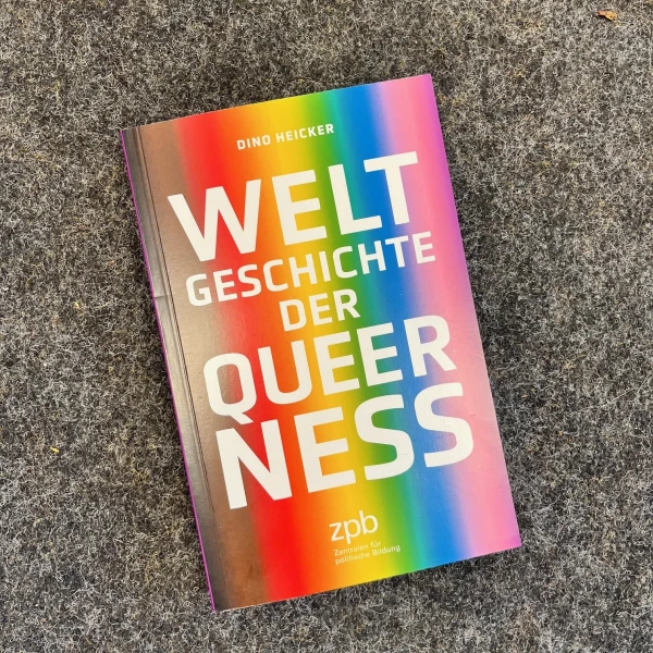 Auf dem Bild liegt ein Buch mit dem Titel "Weltgeschichte der Queerness" auf einem grauen Untergrund. Das Buchcover zeigt einen farbenfrohen Regenbogeneffekt, der von links unten nach rechts oben verläuft. Der Name des Autors "Dirk Wicke" und der Name des Verlags "zpb" sind ebenfalls auf dem Cover zu sehen.  