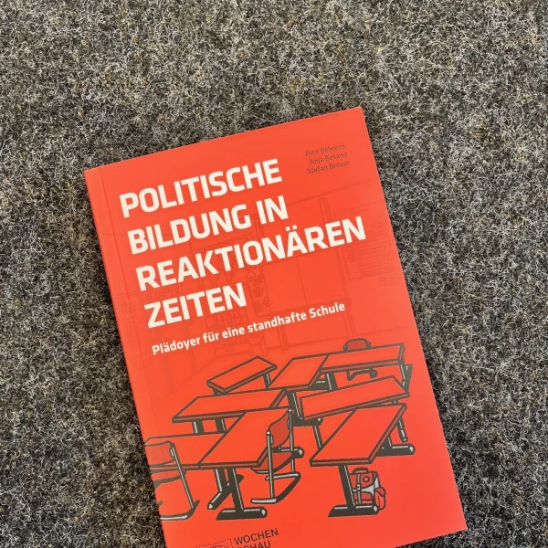 Das Bild zeigt ein Buch mit dem Titel "Politische Bildung in reaktionären Zeiten" in leuchtendem Rot auf dem Cover. Auf der Vorderseite des Buches sind stilisierte, schwarz gezeichnete Figuren abgebildet, die an Schulbänken sitzen und auf einen erhöhten Redner ausgerichtet sind. Das Buch liegt auf einem grauen Teppichboden, der als Hintergrund dient.  