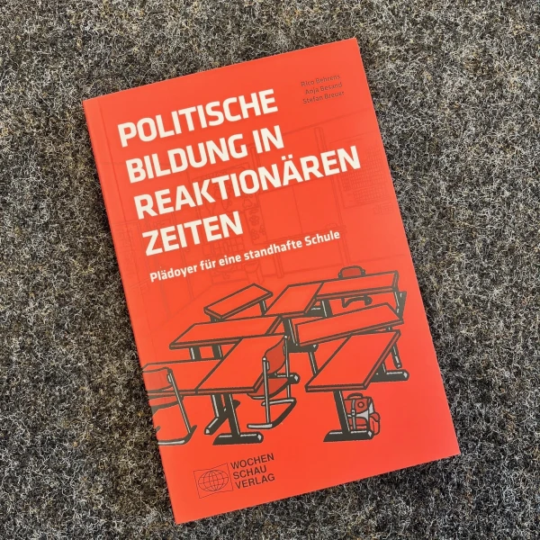 Das Bild zeigt ein Buch mit dem Titel "Politische Bildung in reaktionären Zeiten" in leuchtendem Rot auf dem Cover. Auf der Vorderseite des Buches sind stilisierte, schwarz gezeichnete Figuren abgebildet, die an Schulbänken sitzen und auf einen erhöhten Redner ausgerichtet sind. Das Buch liegt auf einem grauen Teppichboden, der als Hintergrund dient.  