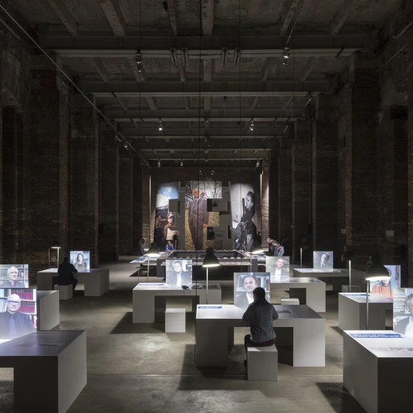 Das Bild zeigt eine Kunstinstallation in einem großen, dunklen Raum mit sichtbaren Backsteinwänden und hohen Decken, betont durch gedimmte Beleuchtung. In der Mitte des Raums sind auf weißen Podesten mehrere leuchtende Displays verteilt, die Porträts und Informationen zeigen. Einzelne Besucher betrachten die Exponate, wobei ihre Aufmerksamkeit auf die beleuchteten Bilder gerichtet ist.  