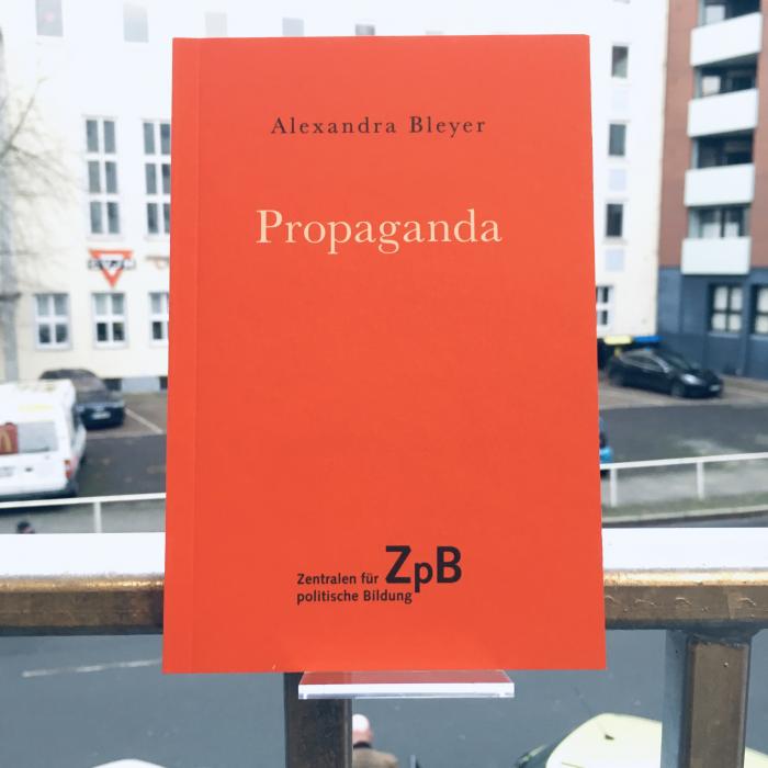 Ein Bild des Buches: "Propaganda" von Alexandra Bleyer mit dem Hintergrund von Gebäuden.