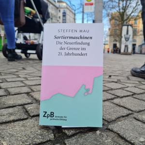 Ein Bild des Buches "Sortiermaschinen-Die Neuerfindung der Grenze im 21. Jahrhundert" von Steffen Mau mit einem Hintergrund einer städtischen Straße.