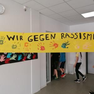 Zu sehen ist ein gelbes Transparent mit schwarzem Text " Wir gegen Rassismus" und Handabdrücken darauf. Im Hintergrund des Plakats laufen ein paar Schulkinder.