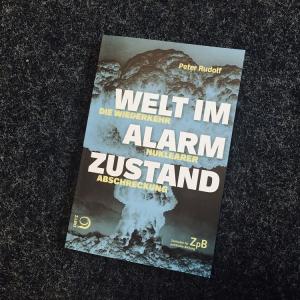 Zu sehen ist Ein Bild des Buchcovers: " Welt im Alarmzustand. Die Wiederkehr nuklearer Abschreckung" mit dem Foto einer Atombombenexplosion. Der Titel auf dem Buch ist weiß und gelb geschrieben.